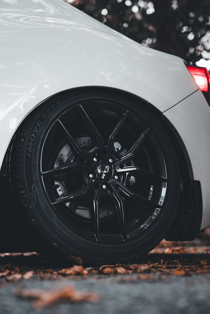 about-img Detailed view of a sporty car's black rim with fallen autumn leaves on the ground.